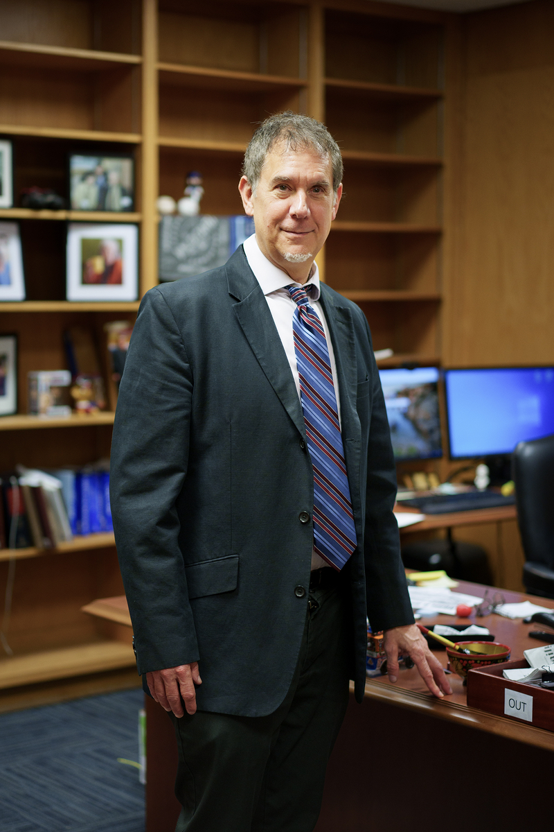 A person in a suit stands beside a cluttered desk in an office, looking at the camera with a slight smile. They wear a striped tie and white shirt under a dark blazer. Behind them are bookshelves with framed photos and personal items, and a computer setup with dual monitors. The desk has various objects including a bowl, a blue mug, and a wooden tray labeled “OUT.”