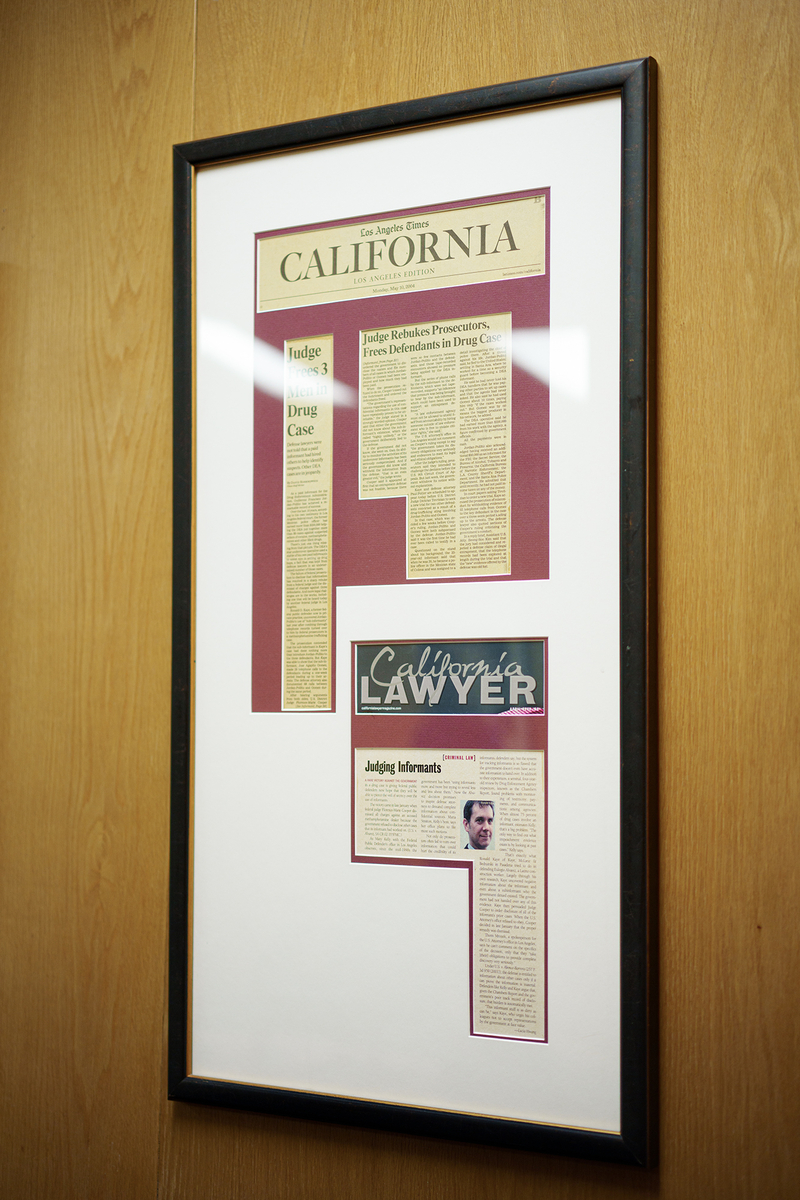 A framed display hangs on a wood-paneled wall, featuring clippings from the Los Angeles Times and California Lawyer magazine. The headlines highlight legal stories, including one titled “Judge Frees 3 Men in Drug Case” and another titled “Judging Informants.” A small portrait of a person is included in the lower article layout. The frame has a dark border and white matting with burgundy backing behind the clippings.