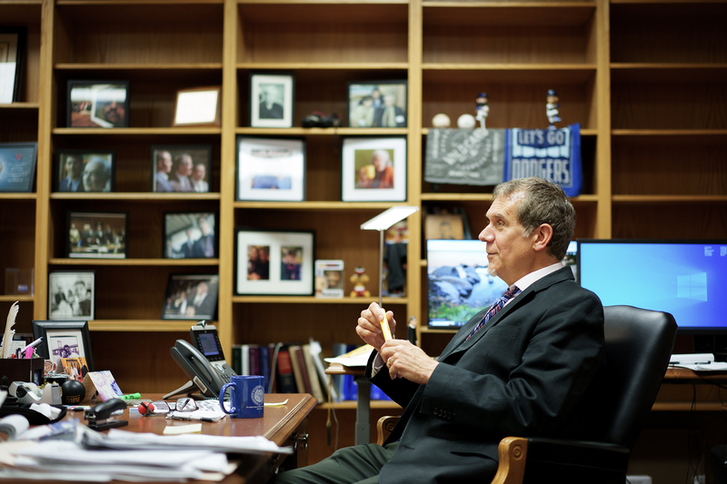 A person in a suit sits in a black office chair behind a cluttered desk, holding a yellow legal pad and pen. Behind them, tall wooden bookshelves are filled with framed photos, books, and sports memorabilia, including a “Let’s Go Dodgers” towel. Two computer monitors are on the desk, one displaying a desktop screen and the other showing a nature wallpaper.