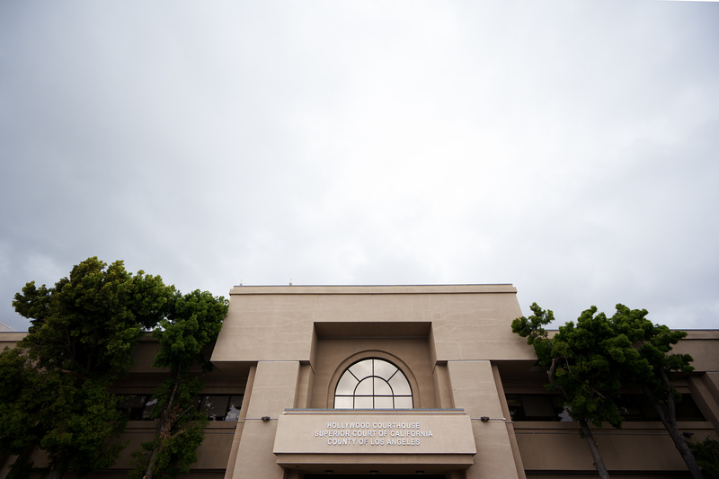 The exterior of the Hollywood Courthouse, a beige concrete building with a large arched window above the entrance. The sign reads “Hollywood Courthouse, Superior Court of California, County of Los Angeles.” Tall trees frame both sides of the building, and the sky above is overcast.