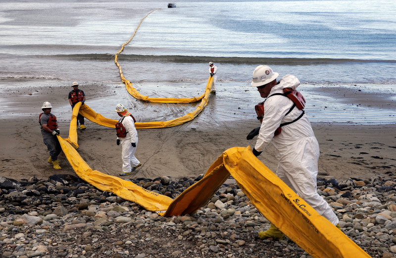 Cleanup workers in protective gear and life vests deploy a long yellow containment boom on a beach following an oil spill. The boom stretches from the rocky shore into the water, with a boat visible in the distance.
