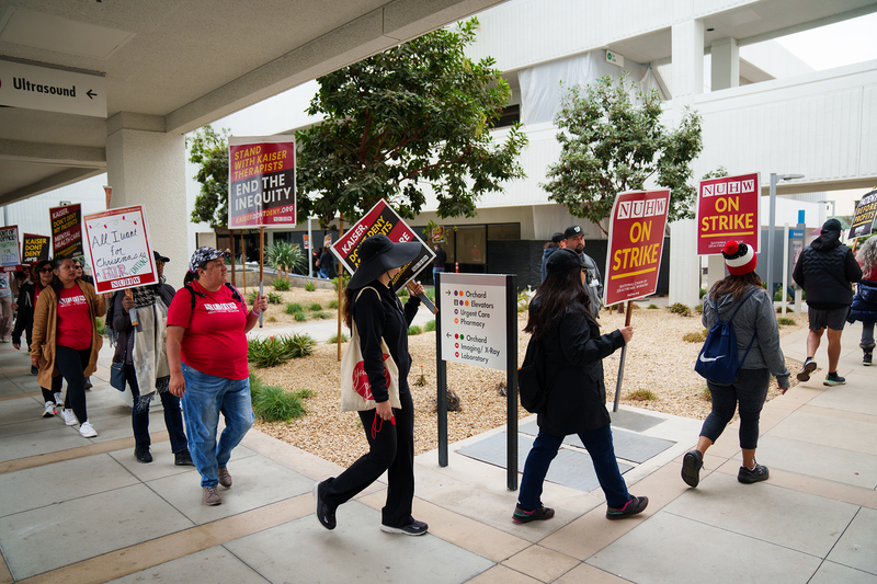 People walking one behind the other turn a corner while holding signs that read "NUHW On Strike" while protesting at a medical building.