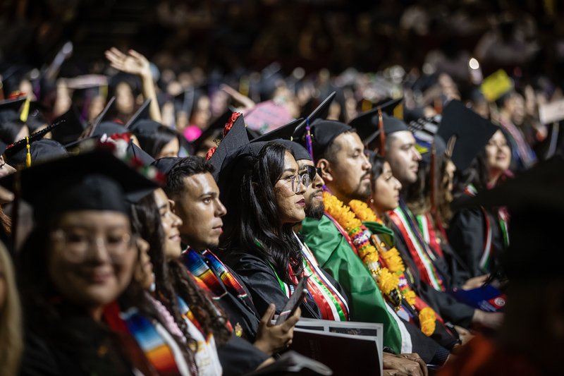 A group of college graduates sits in rows during a commencement ceremony, wearing black caps and gowns adorned with colorful stoles, leis, and sashes. The focus is on a young woman in glasses looking ahead, surrounded by classmates with celebratory decorations, including a student in a green gown and marigold leis.