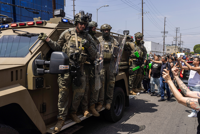 A group of heavily armed law enforcement officers in tactical gear ride on the side of an armored vehicle during a protest. They carry riot shields and crowd-control weapons with green markings. On the right, protesters confront them, shouting, recording on phones, and raising their hands in defiance. The scene takes place on a city street in broad daylight.