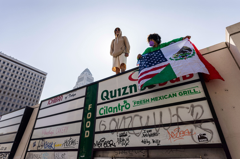 Two people stand on top of a concrete wall with signs of local businesses nearby during a protest. One person holds an U.S.-Mexico flag.