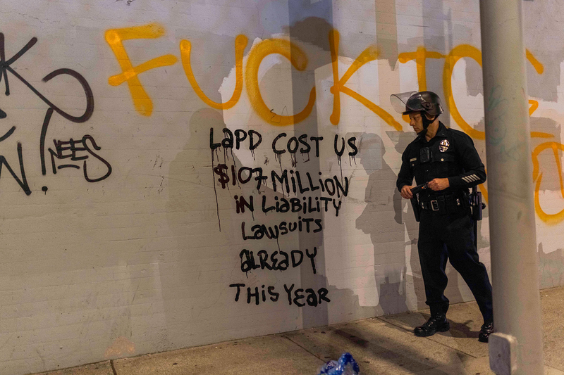 A law enforcement officer looks at graffiti on a concrete wall that says "LAPD Costs Us $107 million in liability lawsuits already this year."