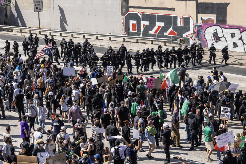 A large crowd of protesters holding signs and flags, including Mexican and American flags, gathers on a street during a demonstration. Opposite them, a line of law enforcement officers in full riot gear stands shoulder-to-shoulder, forming a barrier. Graffiti covers the walls behind the officers, and the scene is marked by tension and confrontation.
