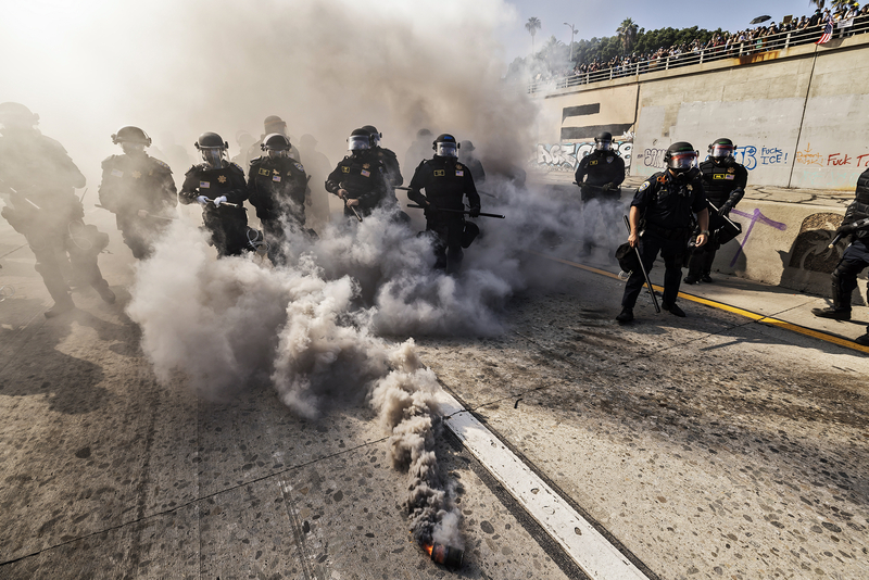 A line of law enforcement officers in full riot gear advances through a cloud of thick smoke on a city street. One smoke canister lies in the foreground, emitting dense gray plumes. Graffiti and a crowd of onlookers can be seen on an overpass in the background.
