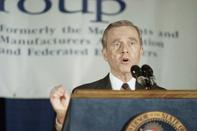 A man in a suit and tie speaks into two microphones at a podium with the seal of the State of California. He gestures with one hand raised slightly. A blurred banner in the background suggests the event is hosted by a business or trade group.