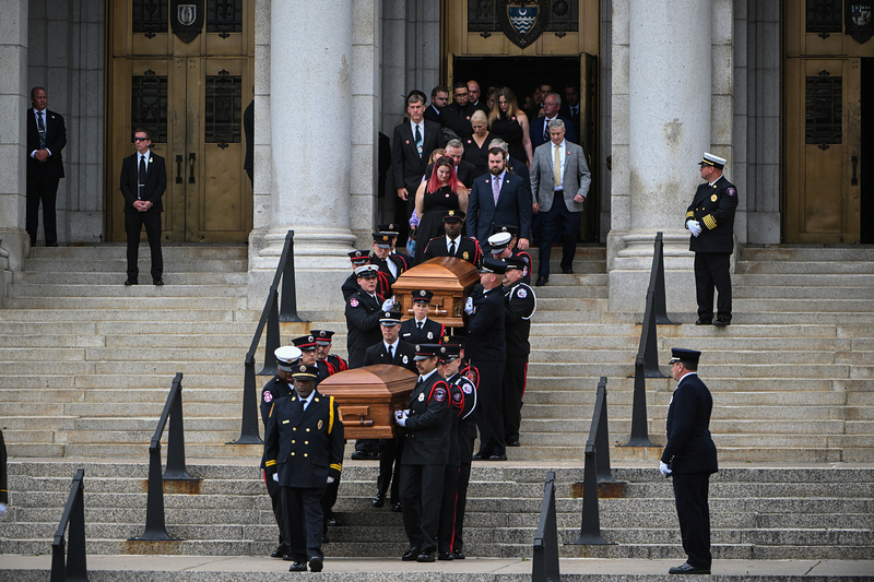 Pallbearers, dressed in formal military uniforms, carry two wooden caskets down a long set of cement steps outside a church as family members and loved ones follow behind.