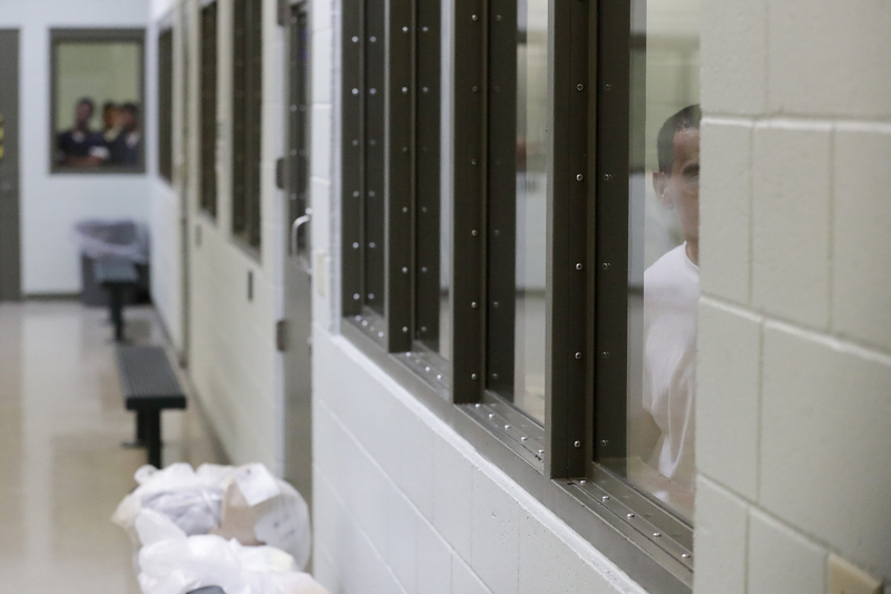 A person stands partially obscured behind a window inside a detention facility. Only half of their face and torso are visible through the reinforced glass. The hallway in the foreground is sterile and institutional, with white walls, benches, and a stack of plastic bags on the floor.