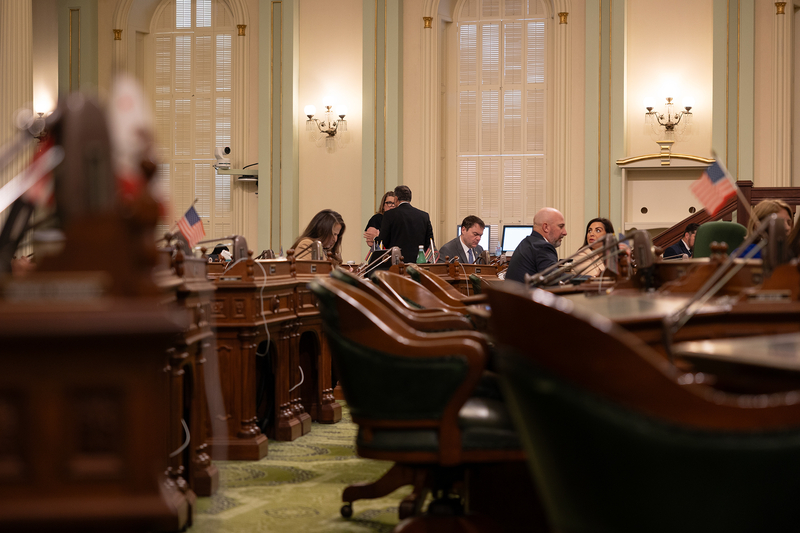 A side view of wooden pedestal desks inside a legislative room adorned with light green and beige features. A couple of lawmakers sit in front of the desks in the far distance.