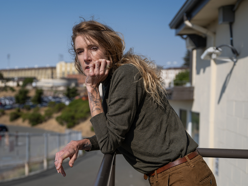 A person with long, windswept hair leans on a railing, gazing directly at the camera with a thoughtful expression. They wear a forest green long-sleeve shirt and brown pants, with visible tattoos on their hand and forearm. The background shows the tan walls and fencing of a prison facility under a clear blue sky, contrasting with the person's relaxed but intense presence in the foreground.