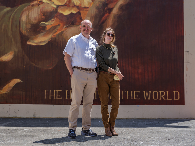 Two people stand side by side in front of a large mural that features glowing yellow flower petals and the words “THE HEART OF THE WORLD.” One person wears a light button-up shirt and khaki pants, smiling with hands in pockets. The other wears a dark long-sleeve shirt, brown pants, and sunglasses, holding a visitor badge and standing casually with one foot turned out. The painted background creates a warm, radiant backdrop behind them.