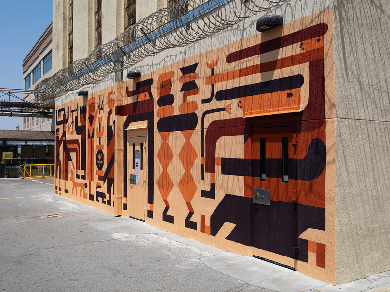 A vibrant geometric mural painted in shades of orange, maroon, and peach covers the exterior wall of a prison building topped with razor wire. The artwork features abstract shapes, figures, and patterns, creating a lively contrast against the institutional architecture and stark concrete surroundings. Two heavy-duty doors are embedded in the mural, blending into the design.