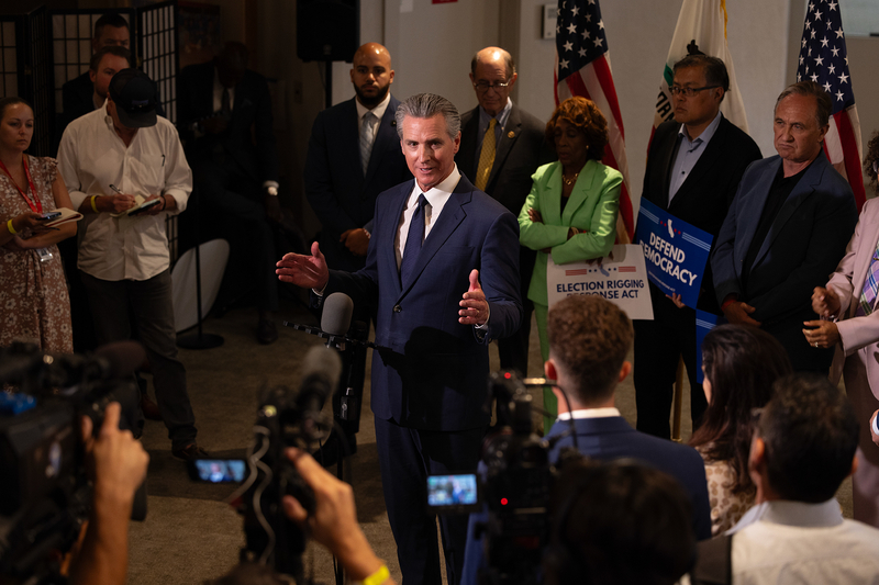 A person, wearing a navy suit, stands in front of a stand with a microphone one it while surrounded by reporters taking notes or filming with cameras during a press conference.