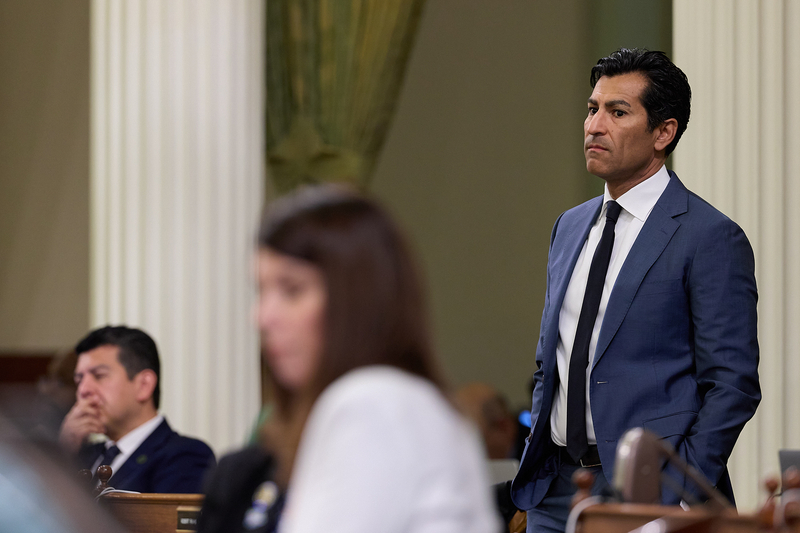 A person, with black slick-back hair and wearing a blue suit, stands with their hands inside their pant pockets during a legislative hearing. Other people can be seen in the background and foreground.