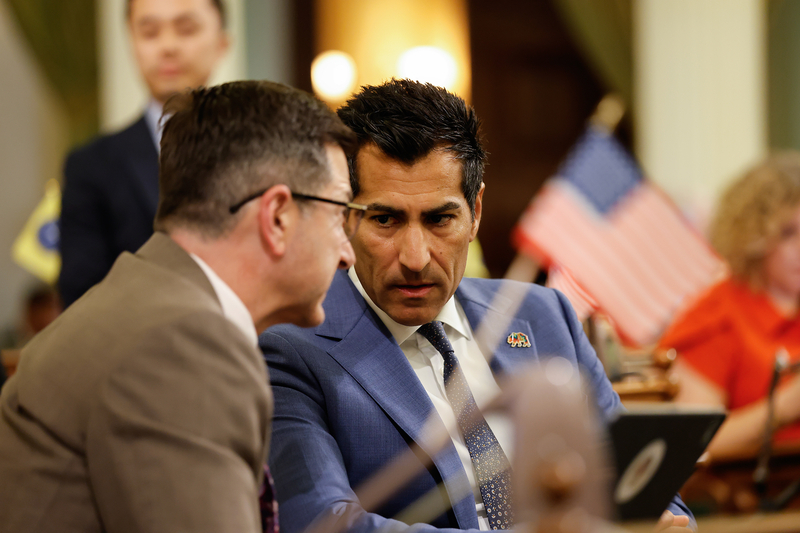 A close-up view of two lawmakers speaking to each other while sitting in front of desks. The lawmaker on the left wears a brown suit, while the lawmaker on the right wears a blue suit. A small and blurred American flag can be seen in the foreground.