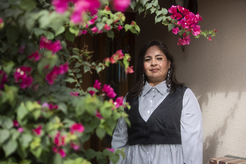 A person wearing a button-down shirt and a vest stands in the front doorway of a home, near a bush of pink and red flowers that peeks into the frame of the photo. Small shadows form on the person's face, created by the branches of the bushes, while the sun shines on their face.