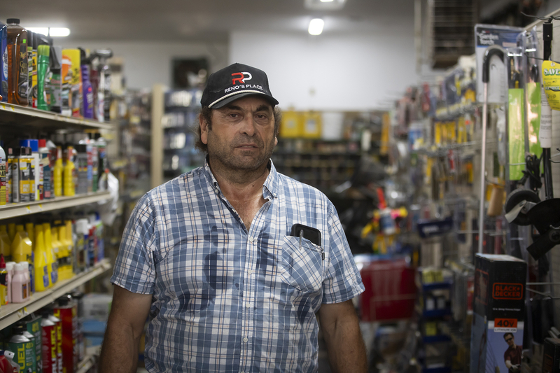 A person wearing a trucker's hat stands in the aisles of a hardware store with fluorescent light shining above and aisles of hardware items for sale on the shelves.