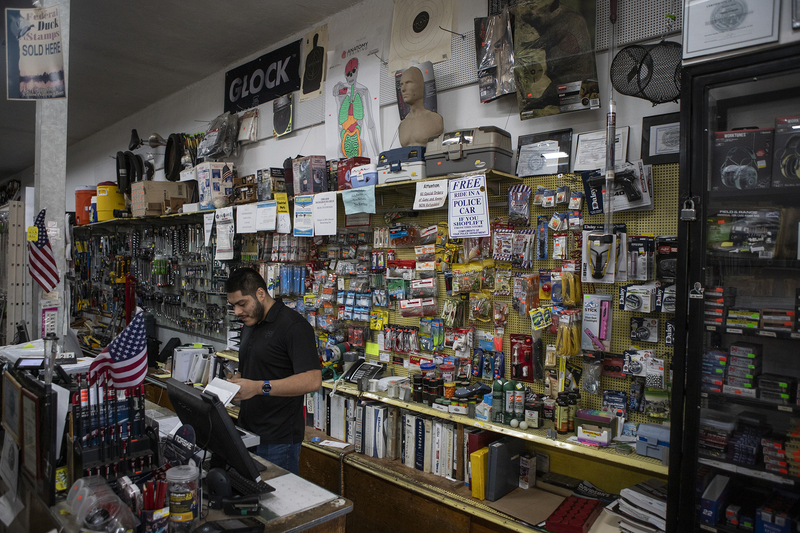 An employee of a hardware store stands at the cash register at the front counter of the store with a busy assortment of hardware items for sale hanging from the wall behind the counter.