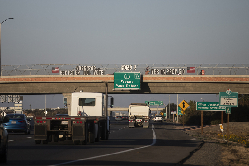 Traffic on a highway drives under an overpass bridge with signs hanging above reading “Worried? Register to Vote,” and another reading “Vote Yes on Prop 50.” Next to the signs are American Flags hanging on the fence and people holding signs.
