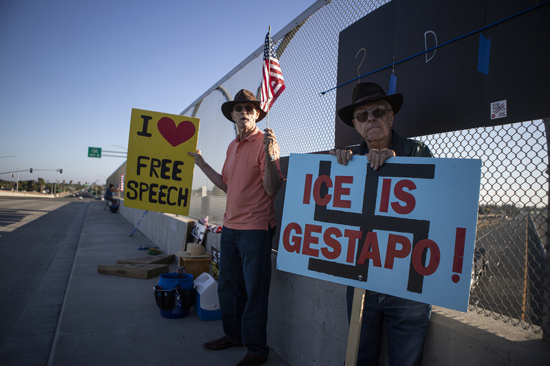 Two protestors stand on the curb side of an overpass holding signs, one saying “I Heart Free Speech,” and the other reading “ICE is Gestapo” as they stand in a shady area with the highway in view behind them.