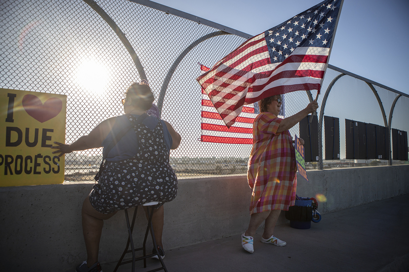 Two protesters stand on an overpass, near a fence, one holding a sign that says “I heart due process,” while the other protester sits, and the first protester waves an American flag in the air as vehicles pass by.
