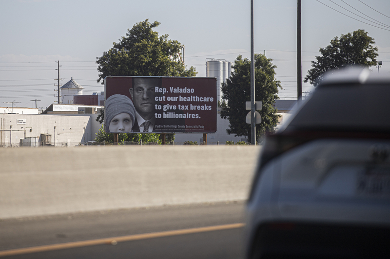 A political sign depicting a politician and a young child reading “Rep. Valadao cut our healthcare to give tax breaks to billionaires” was posted on a billboard near a busy highway, with vehicles passing by at high speeds. The billboard is visible from across the lanes of traffic and over the divider in the middle of the road.