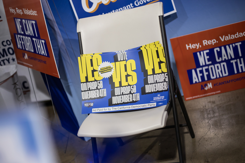 A stack of blue and yellow campaign posters reading “Vote Yes on Prop 50, November 4th” rests on a white folding chair at an event. The posters are surrounded by other political signs, including one criticizing Rep. Valadao, in a crowded booth-like setting.