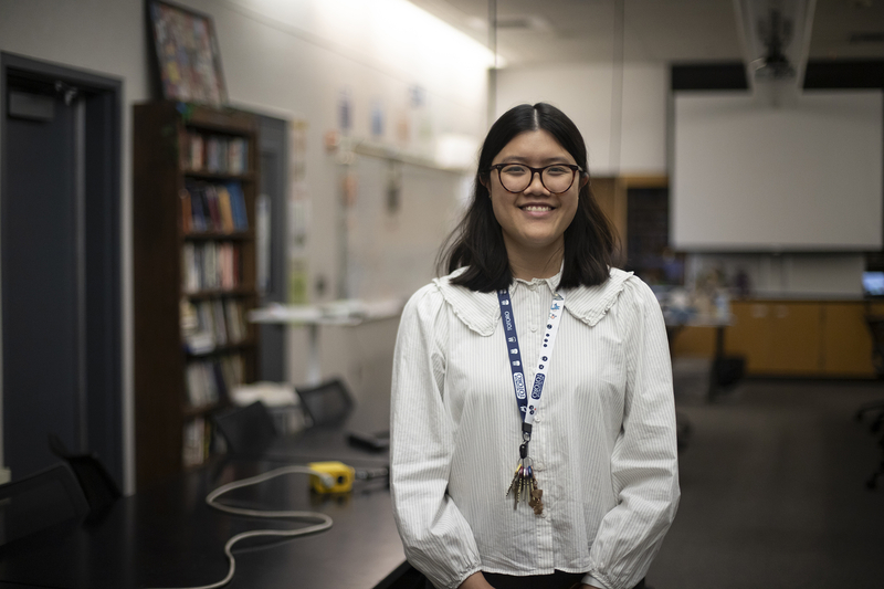 A teacher wearing glasses and a white shirt stands in the center of their classroom with desks, bookshelves, and a projector screen in the background.
