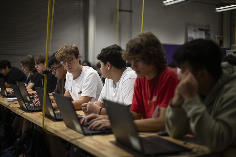 A classroom of students seated at desks in rows with laptops in front of them, talking to each other during a class lesson.