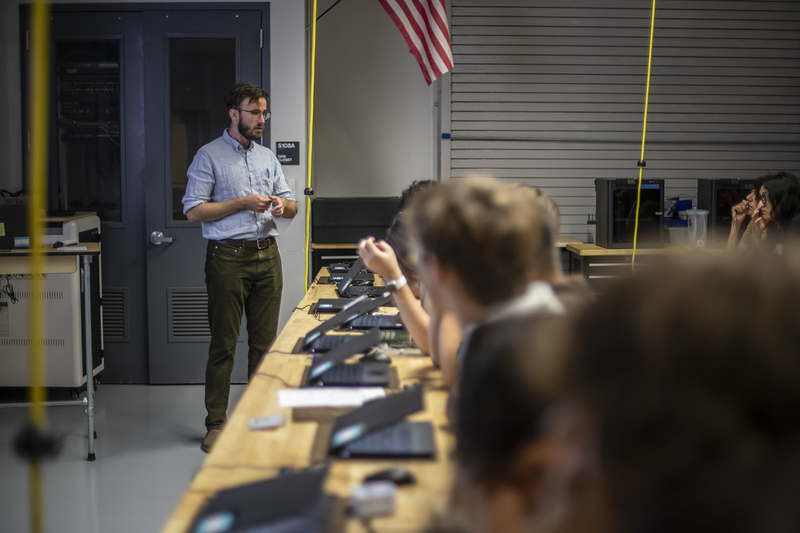 A person wearing glasses stands in front of a classroom of students seated at desks. The students are sitting in rows with laptops in front of them.