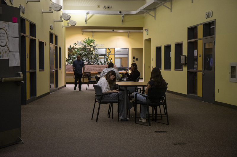 A group of students is sitting at a table set up in the center of a school hallway with saffron-yellow walls and classrooms.
