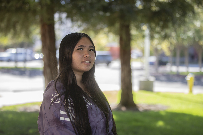 A person with long hair wearing a sweatshirt stands by a group of trees near a parking lot area outside of a school.