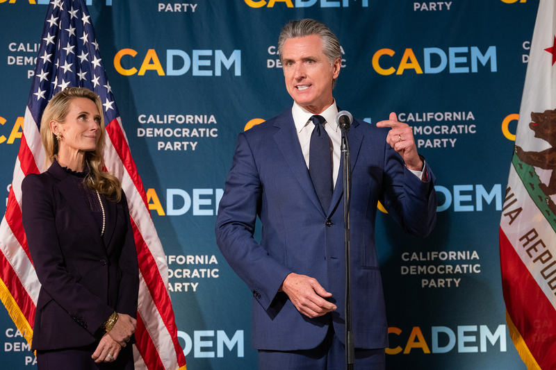 A person, wearing a blue suit, speaks while standing in front of a microphone. Another person, standing on the left and wearing a purple blazer, looks up towards the person in front of the microphone. Both individuals stand in front of a blue step-and-repeat with the California Democratic Party logo. An American flag can be seen nearby.