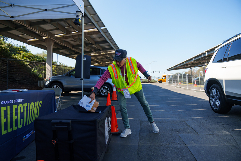 A poll worker, wearing a neon safety vest, places two envelopes into a large black bag as a car sits idle inside a parking lot.