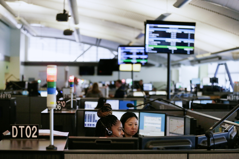 A wide view inside a 911 dispatch center with rows of computer stations and overhead screens displaying call and dispatch information. In the foreground, two dispatchers wearing headsets sit at adjacent monitors, focused on their screens. Color-coded alert lights rise from several desks, and other staff members work in the large, softly lit room.