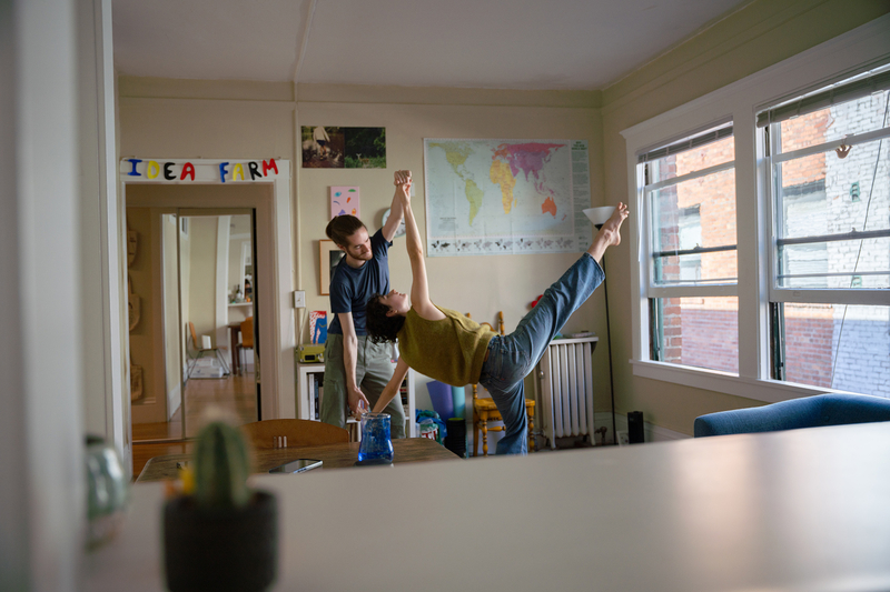 Two people dancing in the center of a decorated room, with one person with their leg spread out into the air and arching in a way, while the other one holds them up.