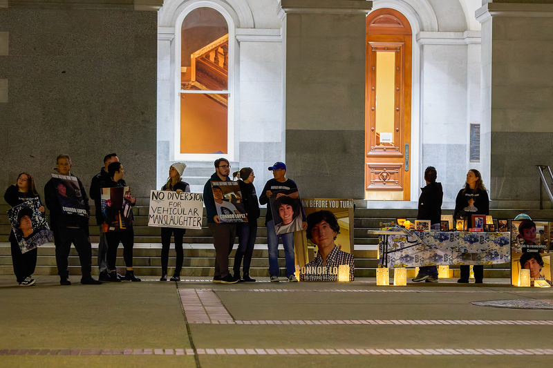 A wide-view of people standing in front of a building while they hold signs and photos of people who have been killed by drunk drivers. One person holds a sign that reads "no diversion for vehicular manslaughter."