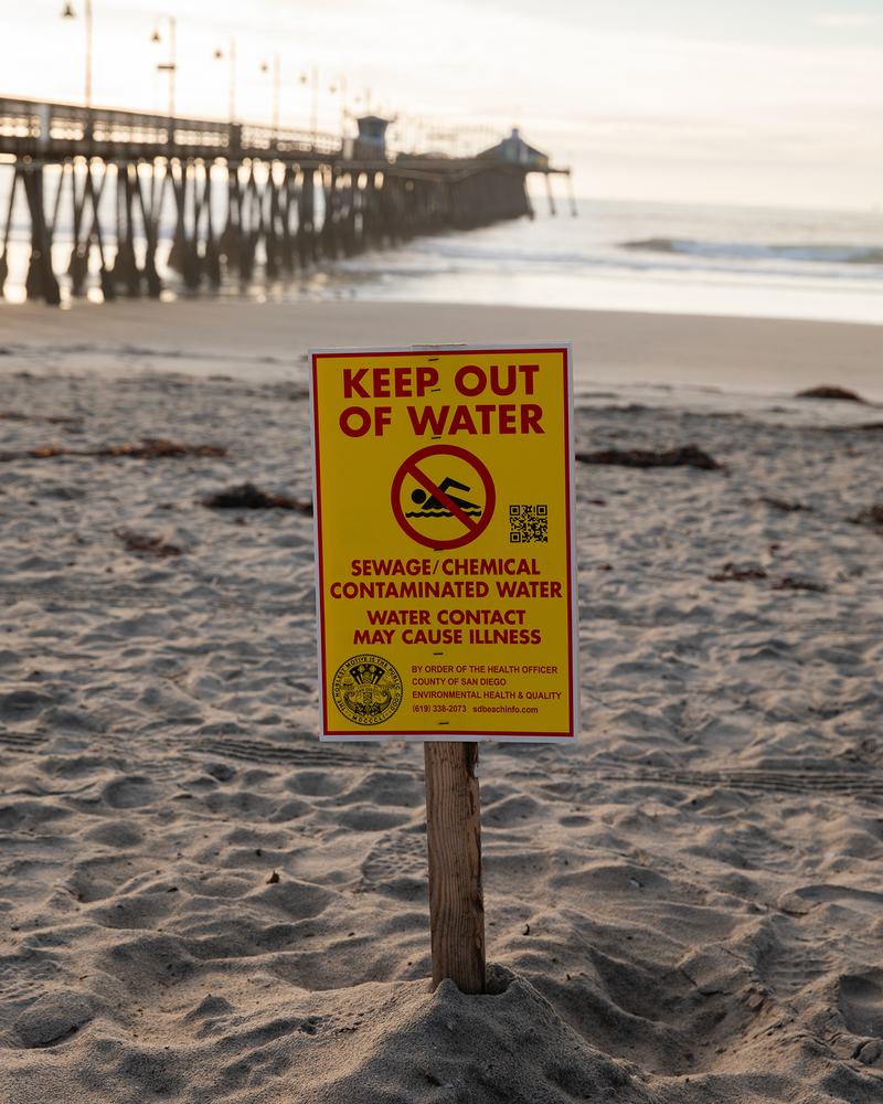 A yellow and red warning sign about sewage and chemical contamination is posted along the shore of a beach. A pier can be seen in the background as the sun begins to set.