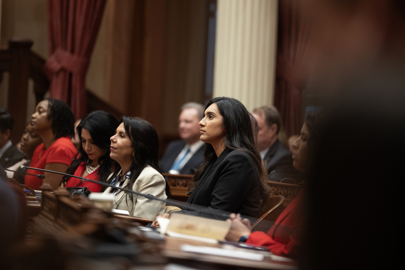 A person wearing a black blazer looks forward as they sit at a wooden desk, surrounded by other people.