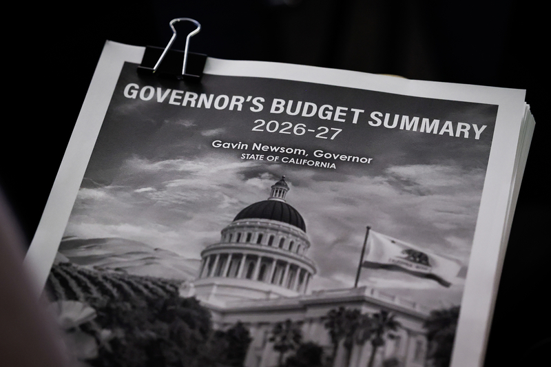 A printed document titled “Governor’s Budget Summary 2026–27” is clipped together, showing a black-and-white image of the California State Capitol with the state flag flying in front.