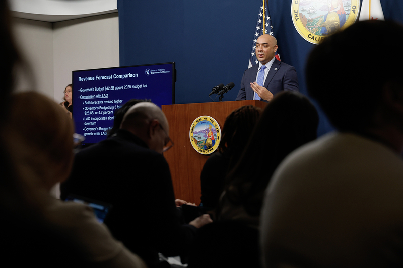 A state official speaks at a podium bearing the California state seal during a budget presentation, with an American flag behind them and a screen displaying a slide titled “Revenue Forecast Comparison,” while members of the audience sit in the foreground.
