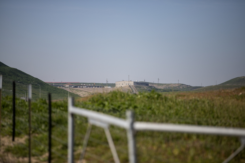 A distant industrial facility sits on a low ridge between rolling green hills, seen beyond a blurred fence and grassy foreground under a pale blue sky.