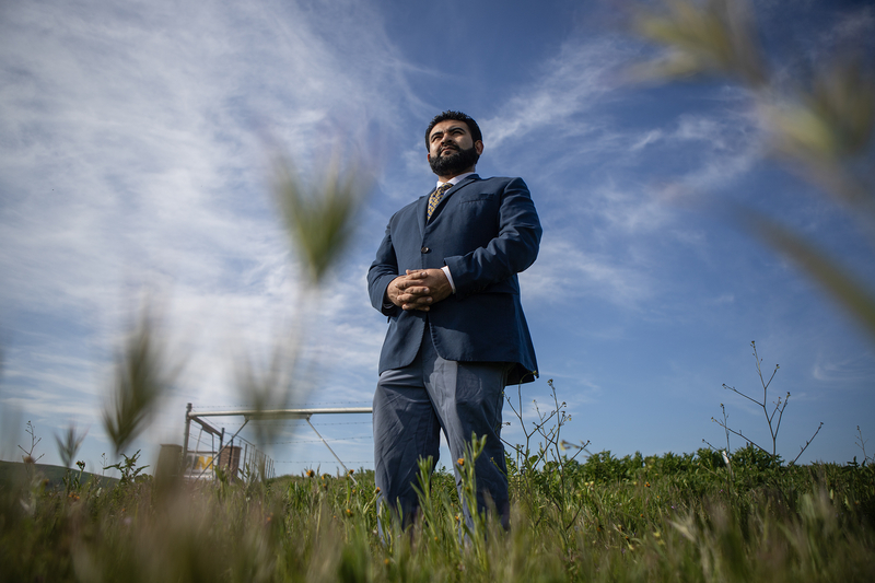 A person in a blue suit stands with hands clasped in a grassy field, photographed from a low angle, with blurred plants in the foreground and a wide sky with wispy clouds overhead.