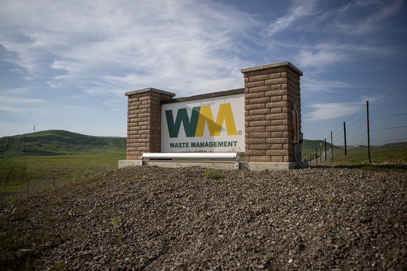 A large Waste Management sign mounted between two brick pillars sits on a gravel slope, with rolling green hills and a partly cloudy blue sky in the background.