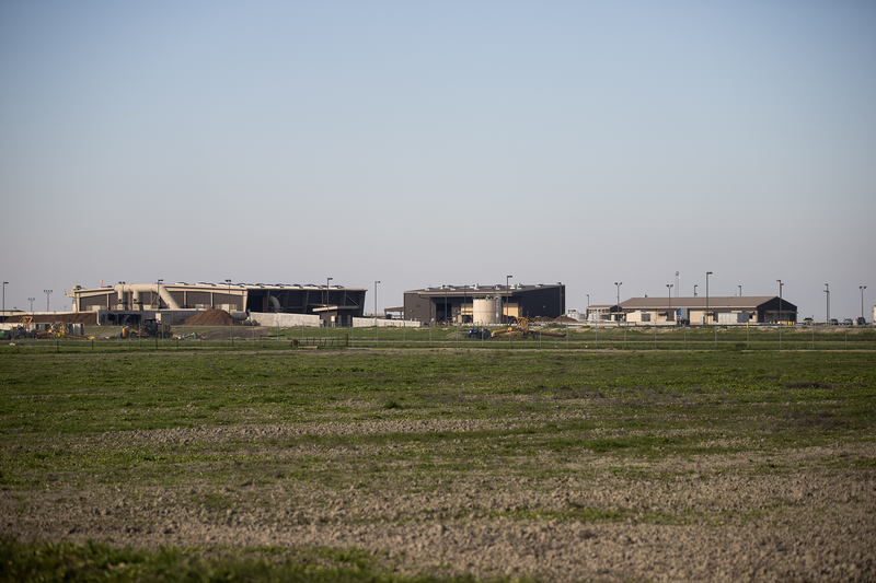An industrial waste management facility stretches across the horizon beyond a fenced field, with large processing buildings, equipment, and service roads visible under a pale blue sky.