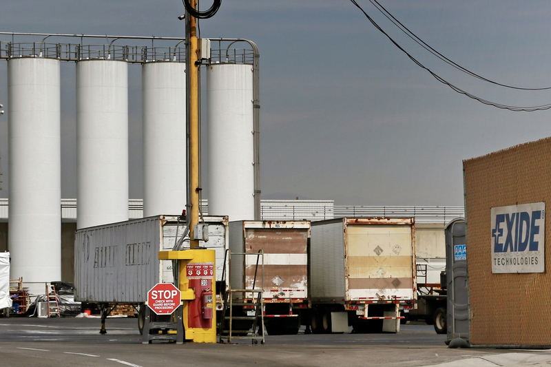 Several semi-trailers are parked in an industrial loading area in front of tall white storage tanks, with a yellow security post and a stop sign in the foreground and an Exide Technologies sign visible on a nearby wall.