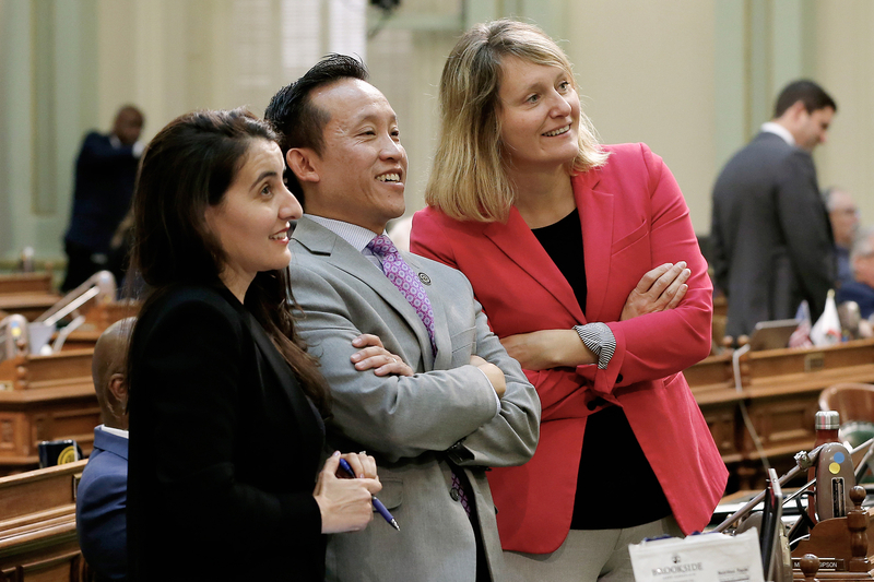Three people stand together inside a legislative chamber, smiling and looking off to one side as if watching something unfold. One person wears a gray suit with a patterned tie, flanked by two others in dark and bright-colored jackets, with desks and other lawmakers visible in the background.